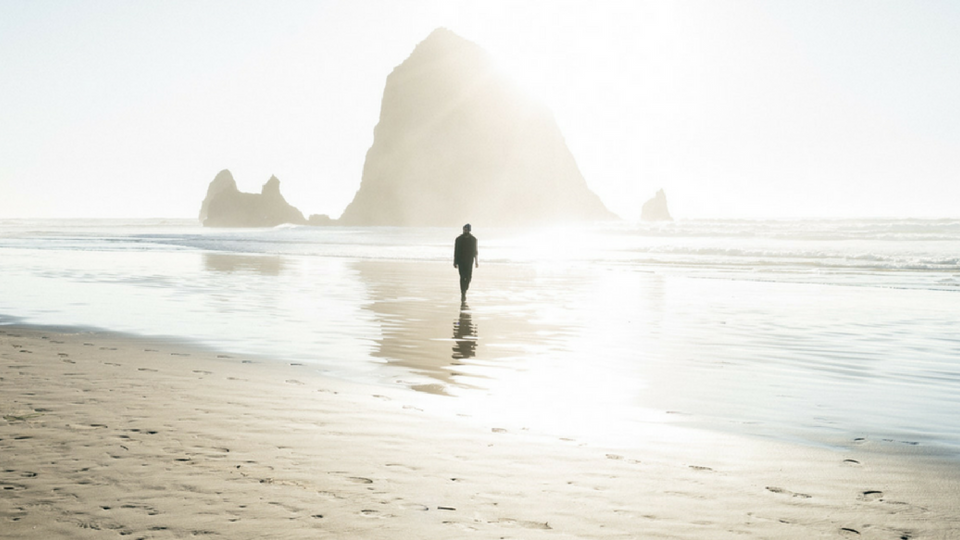 man walking on the beach in the water toward a rock