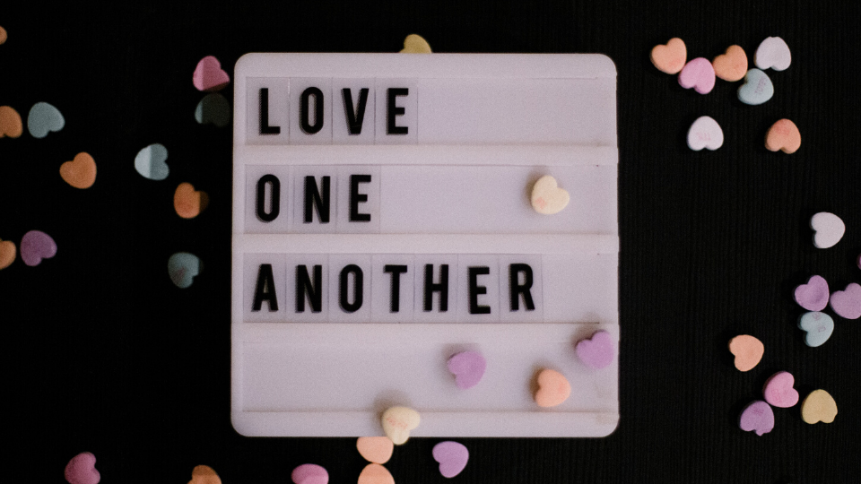 Love One Another spelled out on a letter board surrounded by candy hearts all resting on a black background