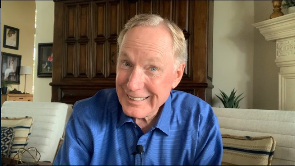 Max Lucado wearing a blue shirt sitting on a white chair with a wooden cabinet behind him