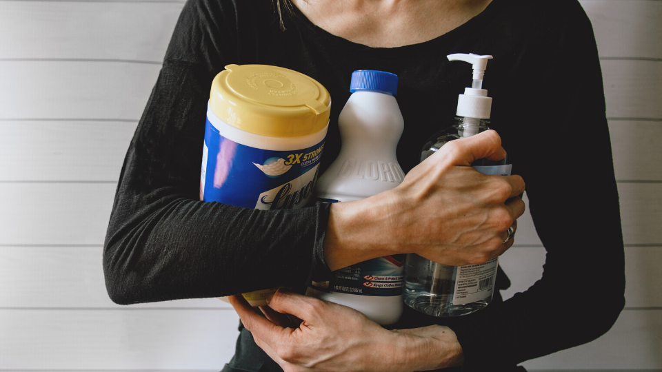 woman wearing a black long-sleeved shirt holding Lysol wipes, bleach, and hand sanitizer