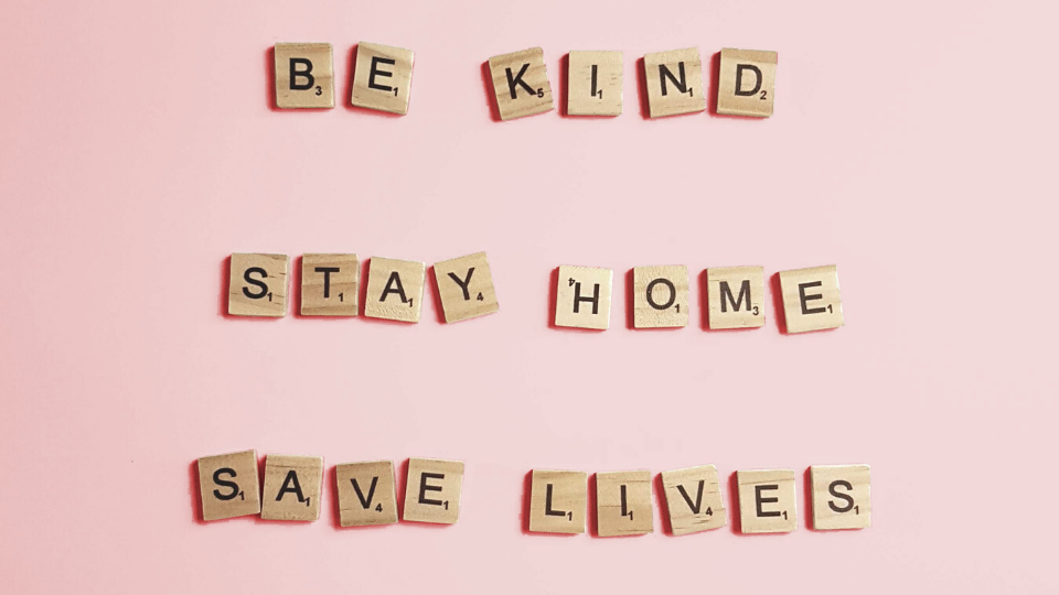 "Be Kind, Stay Home, Save Lives" spelled out in wooden letters on a pink background