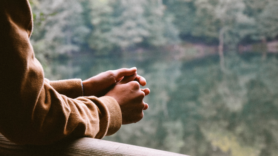 person wearing tan sweatshirt standing on a balcony overlooking a lake with their hands folded