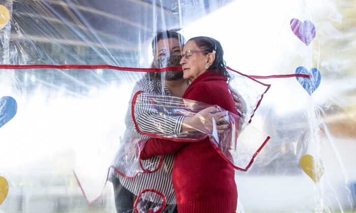 woman in striped shirt hugging elderly woman in red coat through plastic shield
