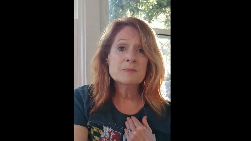 Tracey Tiernan sitting in front of a window wearing a blue shirt and looking into the camera with her left hand on her heart