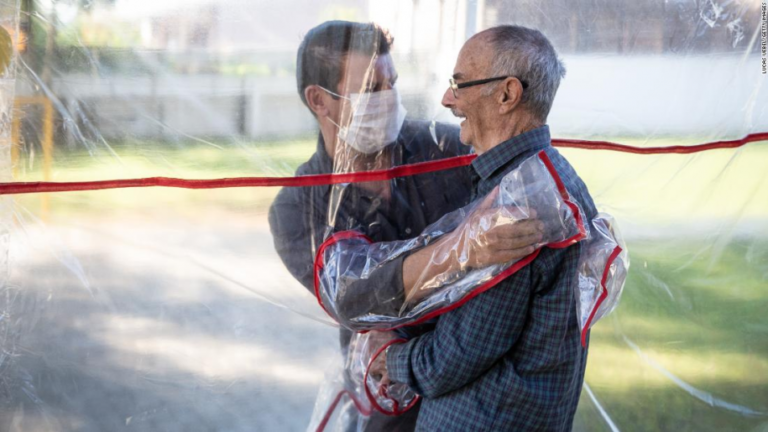 Man and his elderly loved one hugging and looking at each other through plastic shield