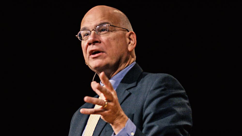 Tim Keller standing and preacher with his left hand extended forward the background is black