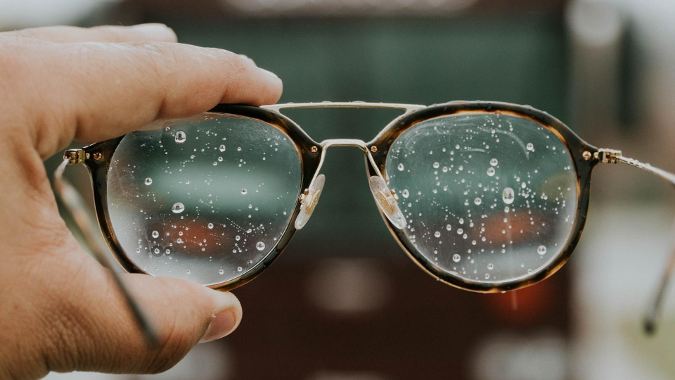 Person holding up glasses in front of camera with water droplets on the lenses