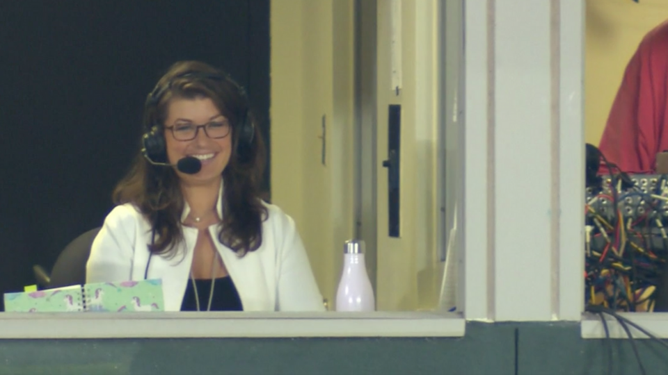 Melanie Newman wearing a white jacket and black headset with microphone smiling from inside a box at Oriole's Camden Yards Park