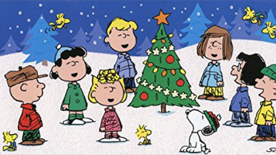 Charlie Brown cartoon and friends standing around green Christmas tree in the snow as they sing