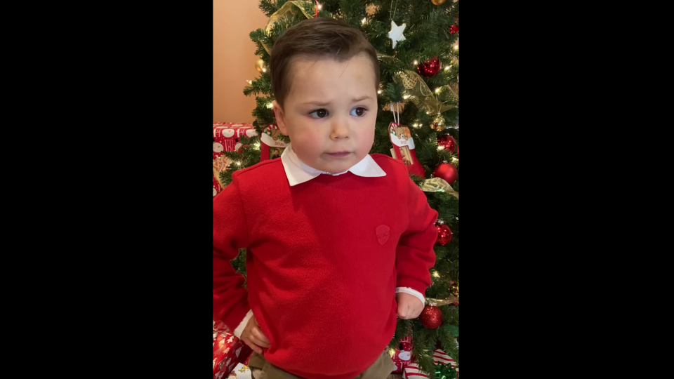 A little boy with brown hair and wearing a bright red sweater with a white collar as he standing in front of a Christmas tree with red ornaments with his hands on his hips