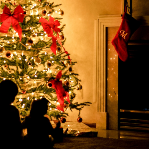 the shadows of two small children in front of a lit up Christmas tree decorated with red bows and ornaments as it sits to the left of a white fireplace