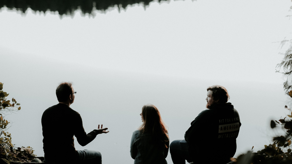 Two men sitting on rocks and a girl sitting in between them the girl and the one man look to their left as a man with glasses talks with his hand out they sit in front of a lake with their backs to the camera