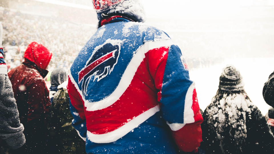 man standing in the stands of a football field wearing a blue and red jacket with a buffalo bills logo on the back as it snows