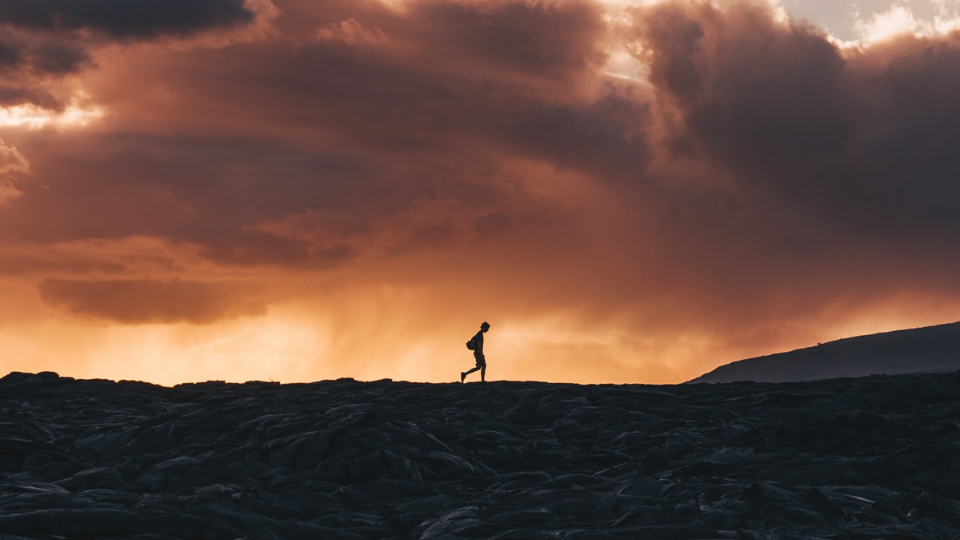 person walking across a plain of black rocks with an orange sky filled with clouds in the background