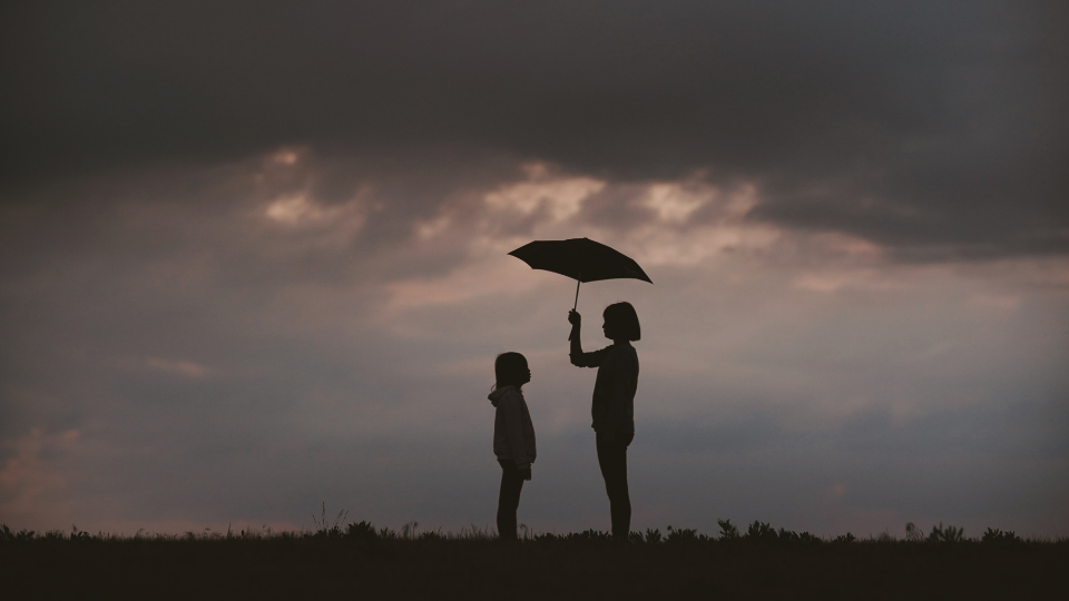 silhouettes of Two children standing face to face while one holds an umbrella over both of their heads underneath a gray stormy sky