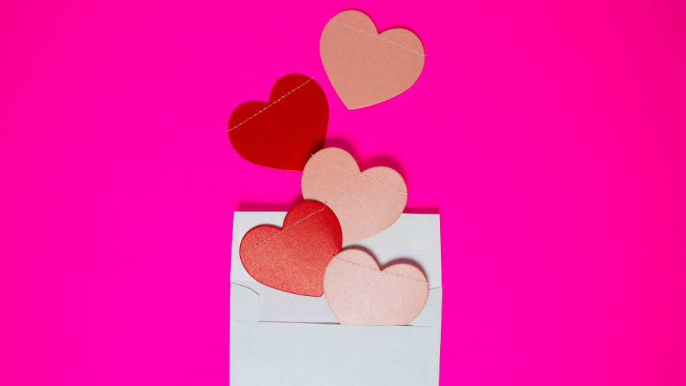 bright pink backdrop with a white envelope laying on it that has red and light pink paper hearts coming out of it