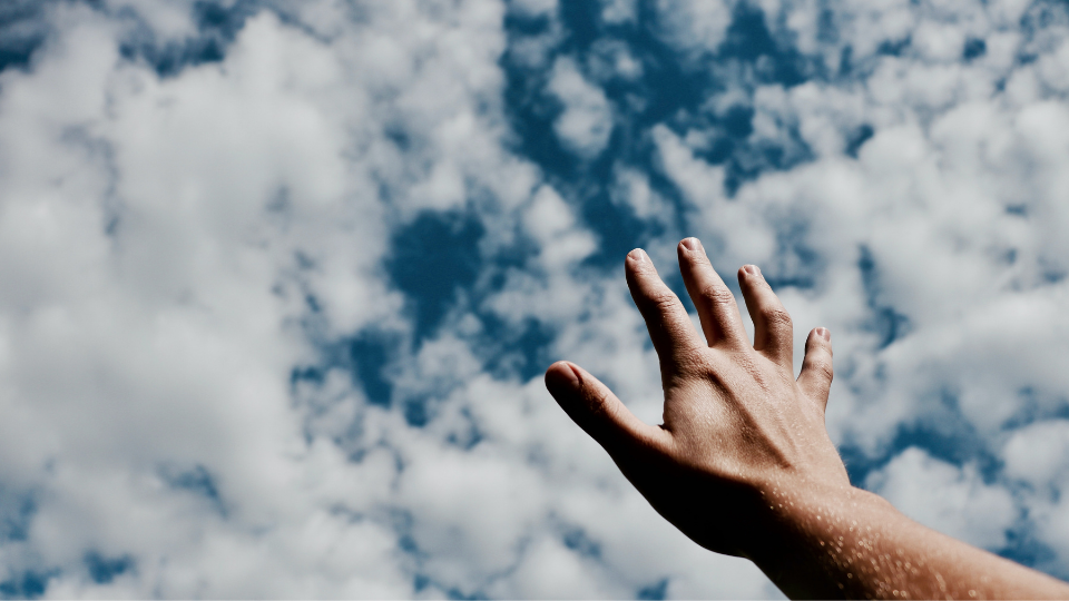person's open hand reaching up towards blue sky with a thin layer of white puffy clouds