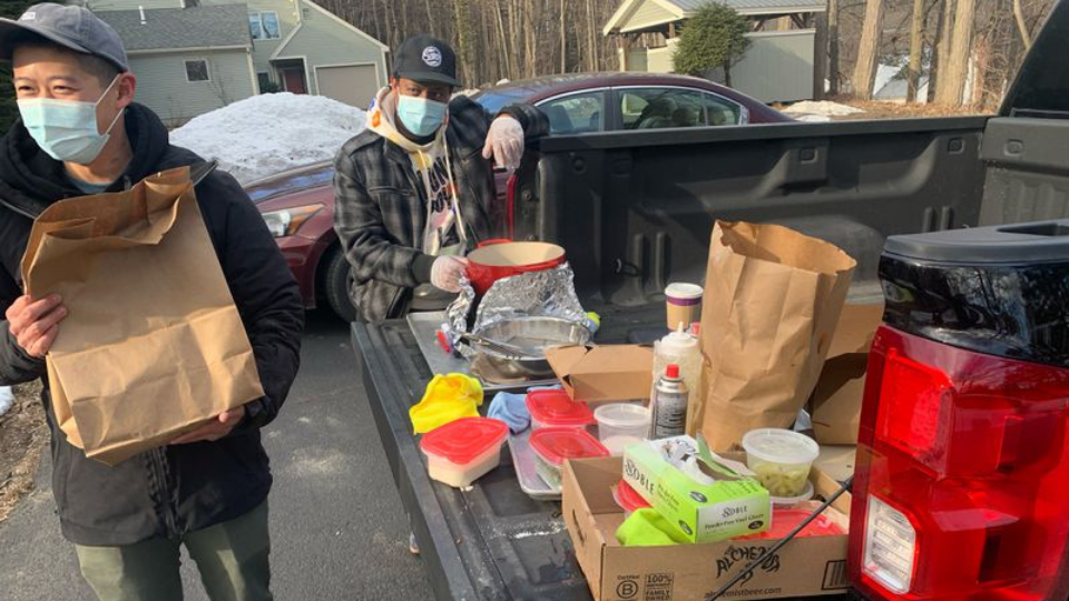 Two men standing beside the back of a pickup truck with food supplies while wearing masks as they prepare to deliver takeout food to terminally ill customer