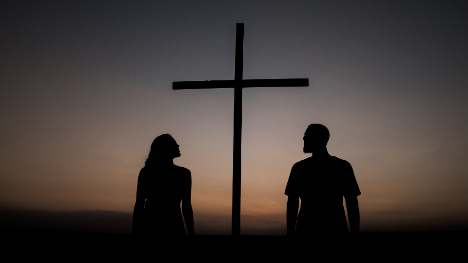 shadow of a man and woman looking up at the wooden cross with the sunset in the background