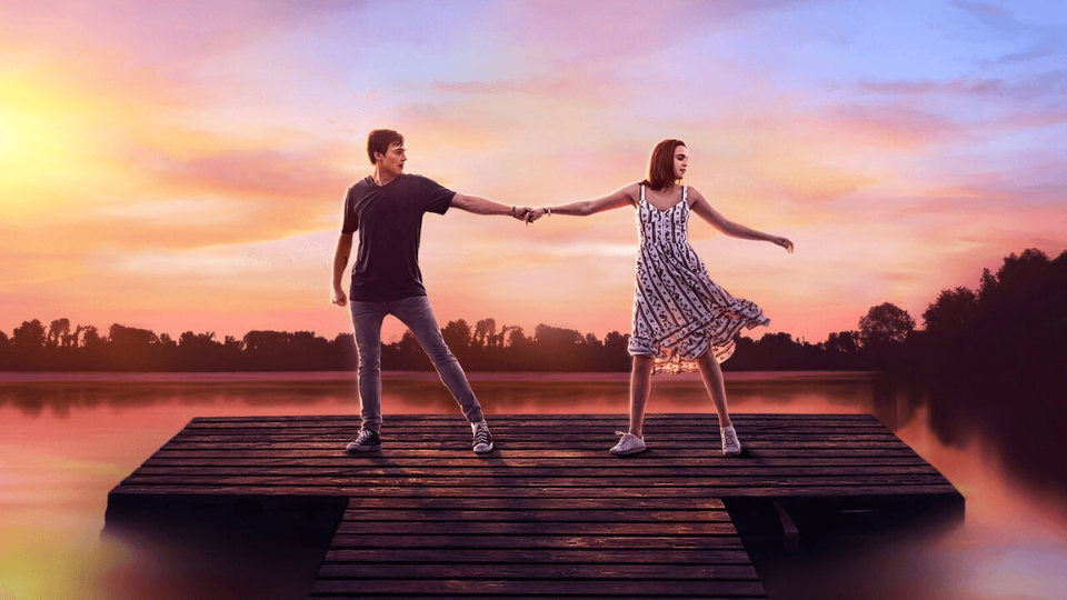 teenage girl and boy holding hands on a pier on a lake as the pink and purple sunset sets in the background