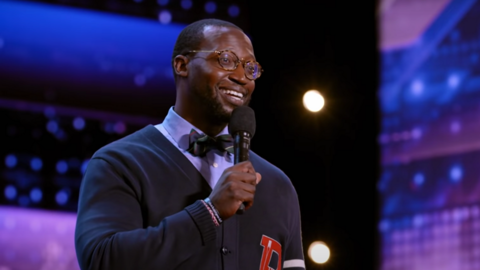 Comedian Mike Goodwin standing on the Americas Got Talent stage smiling while holding a microphone up to his mouth and wearing glasses and a blue button up sweater and a bow tie