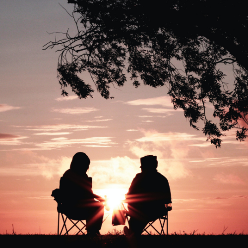 black silhouettes of two people wearing coats and hoods sitting in collapsable lawn chairs watching a pink sunrise near a large tree