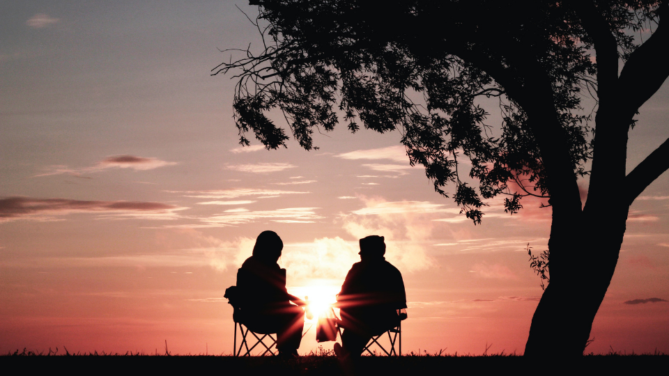 black silhouettes of two people wearing coats and hoods sitting in collapsable lawn chairs watching a pink sunrise near a large tree