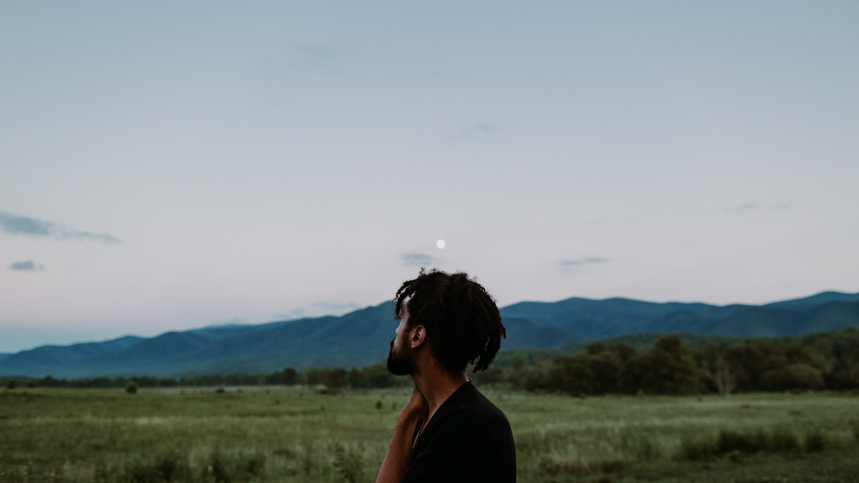 man standing in a green field looking up towards the pale blue sky and mountains as a full moon hangs in above mountain