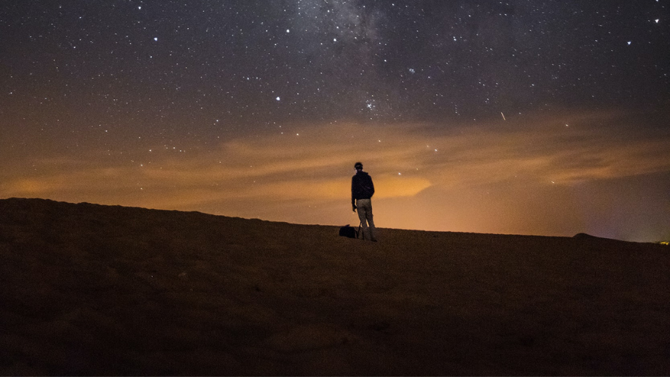 Man standing in a desert looking up a blue and orange sky full of stars