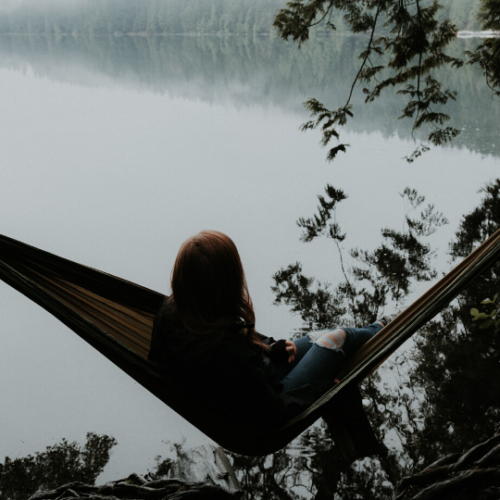 person sitting in a hammock overlooking a still lake while surrounded by leafy trees