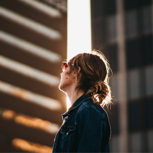woman with low bun and glasses looking up at sun peaking between two city buildings
