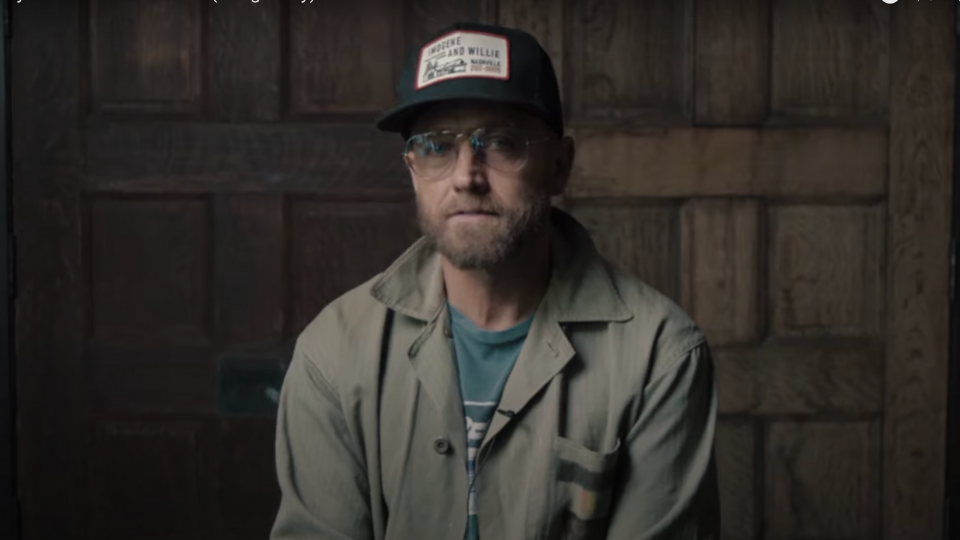 Toby Mac wearing a tan jacket and a black trucker hat sitting in front of set of wooden doors