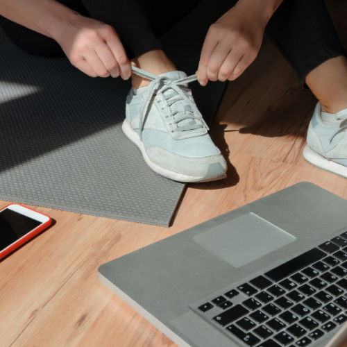 person tying their grey tennis shoes while sitting on a grey yoga mat in front of a laptop on the floor