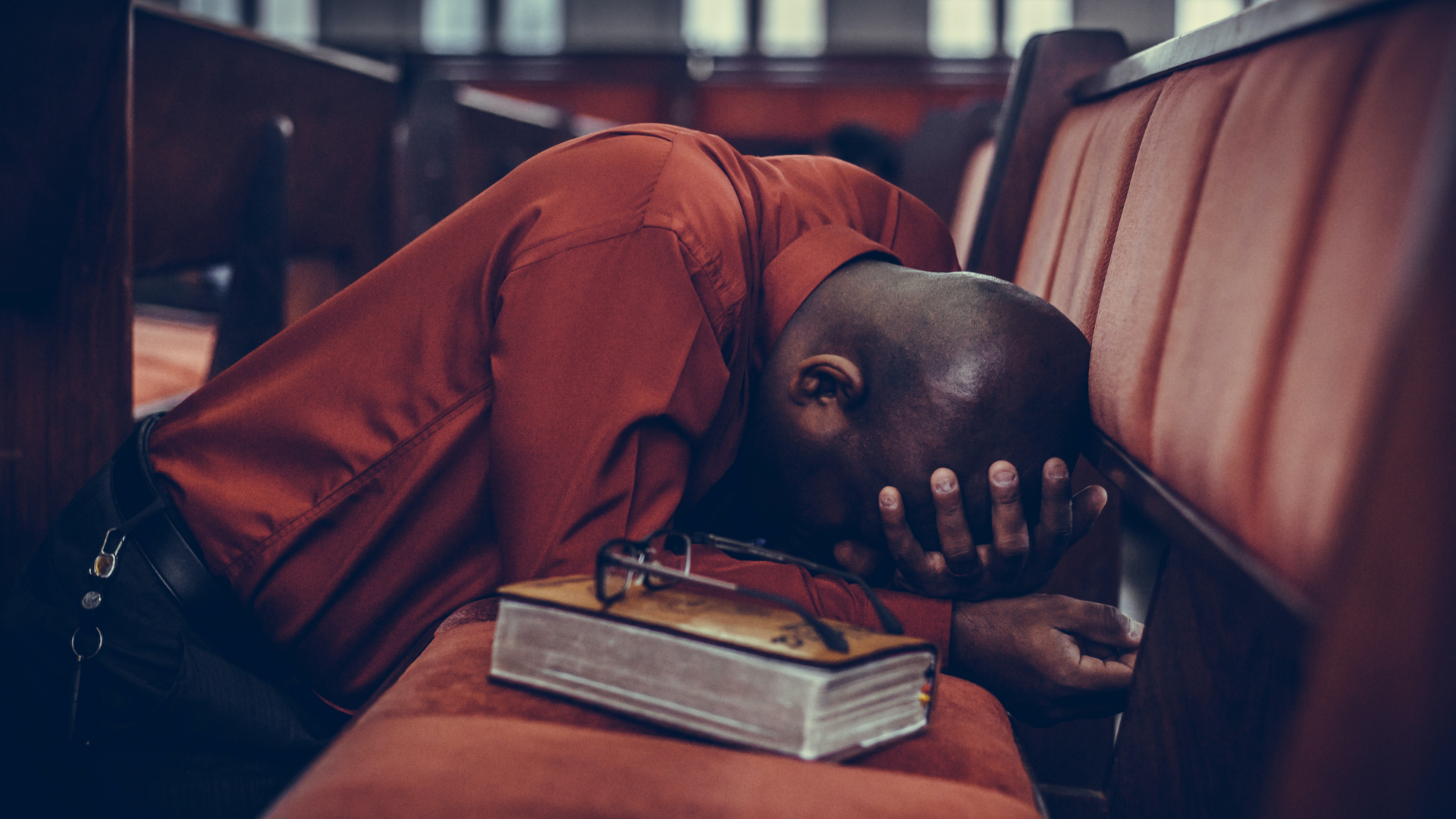 Black man kneeling down in a red church pew holding his head with a Bible and pair of glasses sitting beside him