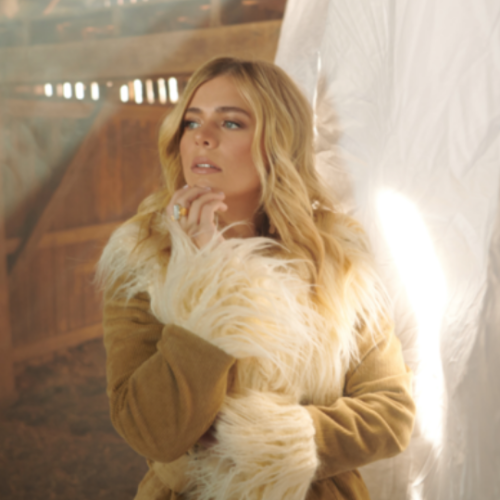 Anne Wilson wearing a tan coat with white fur on the collar standing in a barn in front of a white sheet as the sun shines through