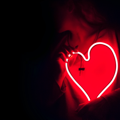 person in a black sweatshirt holding a red neon heart that is lit up