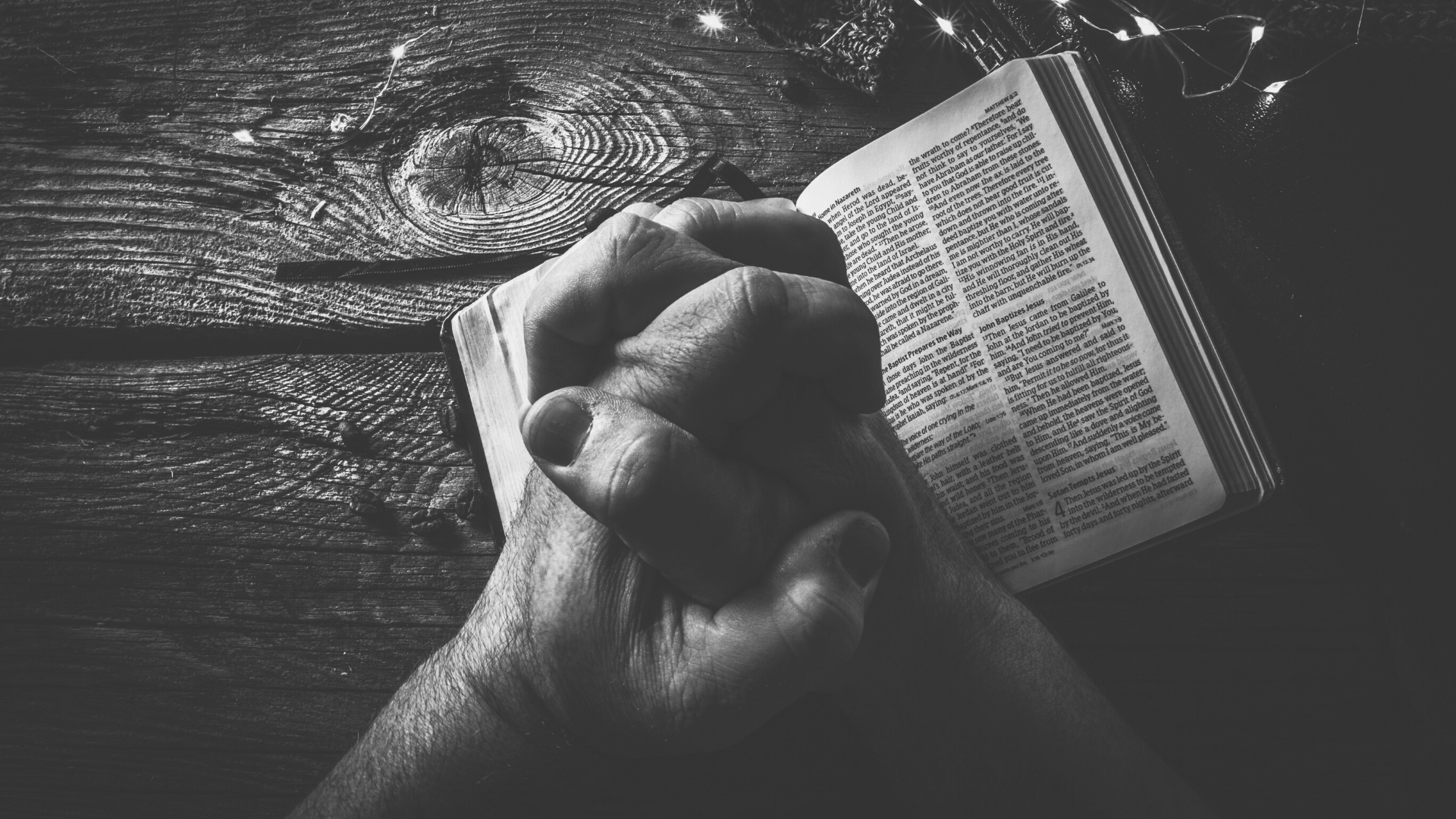 black and white photo of hands clasped together praying over a Bible that it set on a wooden table