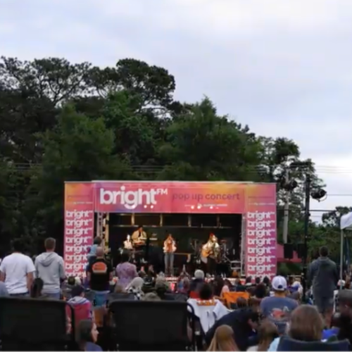 Outdoor concert with people sitting and standing in the grass and a pink and purple and orange BRIGHT FM stage set up