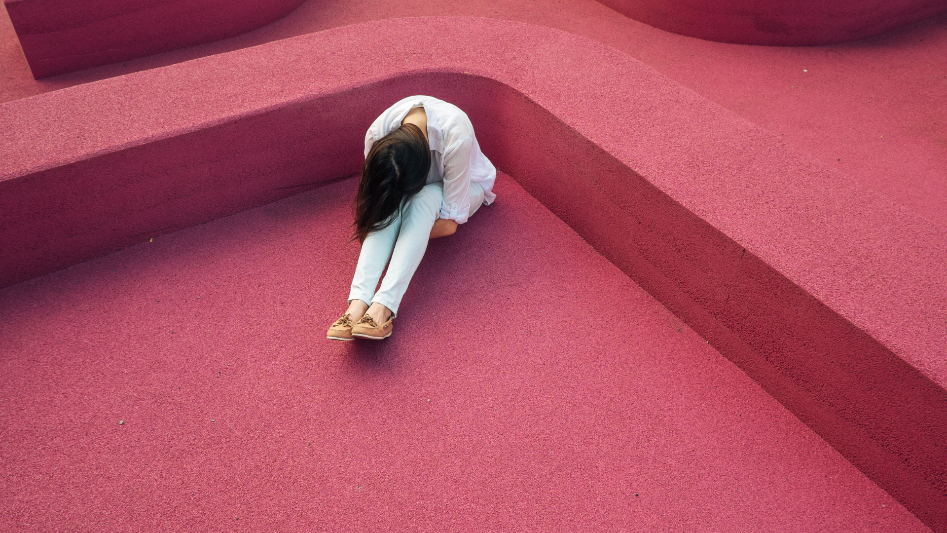 woman with long brown hair sitting on pink pavement with her head between her knees
