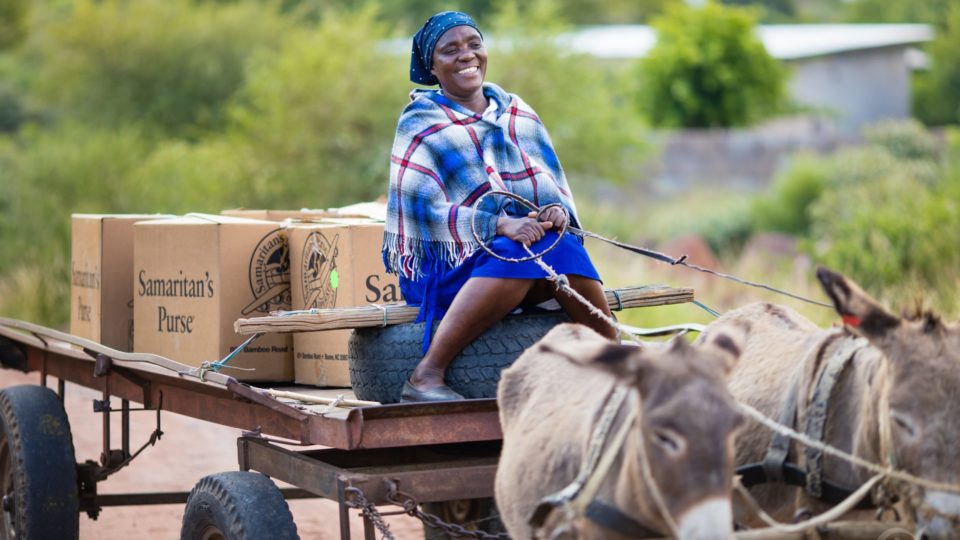 A woman in Botswana sits on a cart stacked with Samaritan's Purse cardboard boxes full of gifts as it is pulled by donkeys