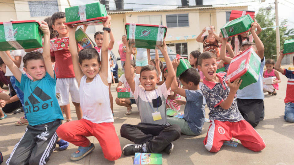 Boys in Colombia sitting on the ground, smiling and laughing as their hold their shoeboxes high above their heads