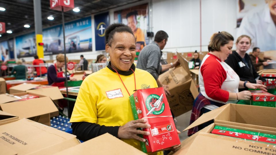 Pictured here is a volunteer smiling and holding up a shoebox before it is packed and sent off to its destination.