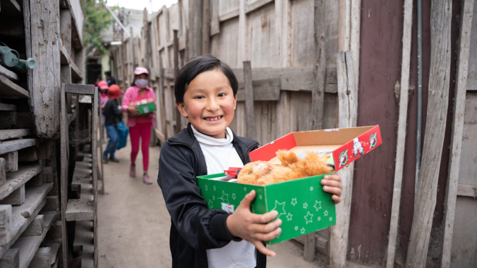 A child in Ecuador standing in a wooden doorway and smiling while holding their open shoebox.