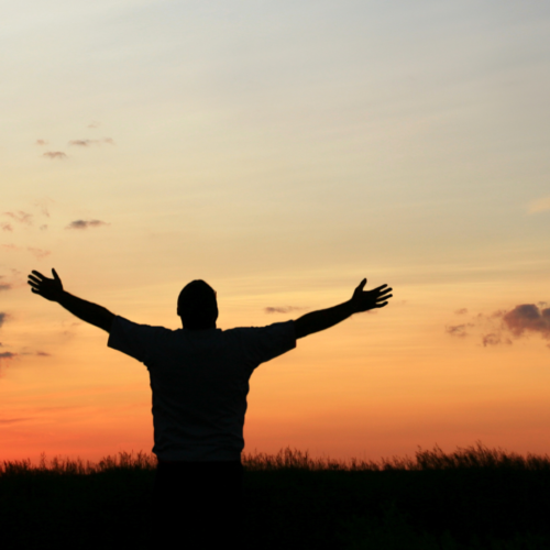 person standing in front of an orange sunset with their arms stretched out and up in worship of God