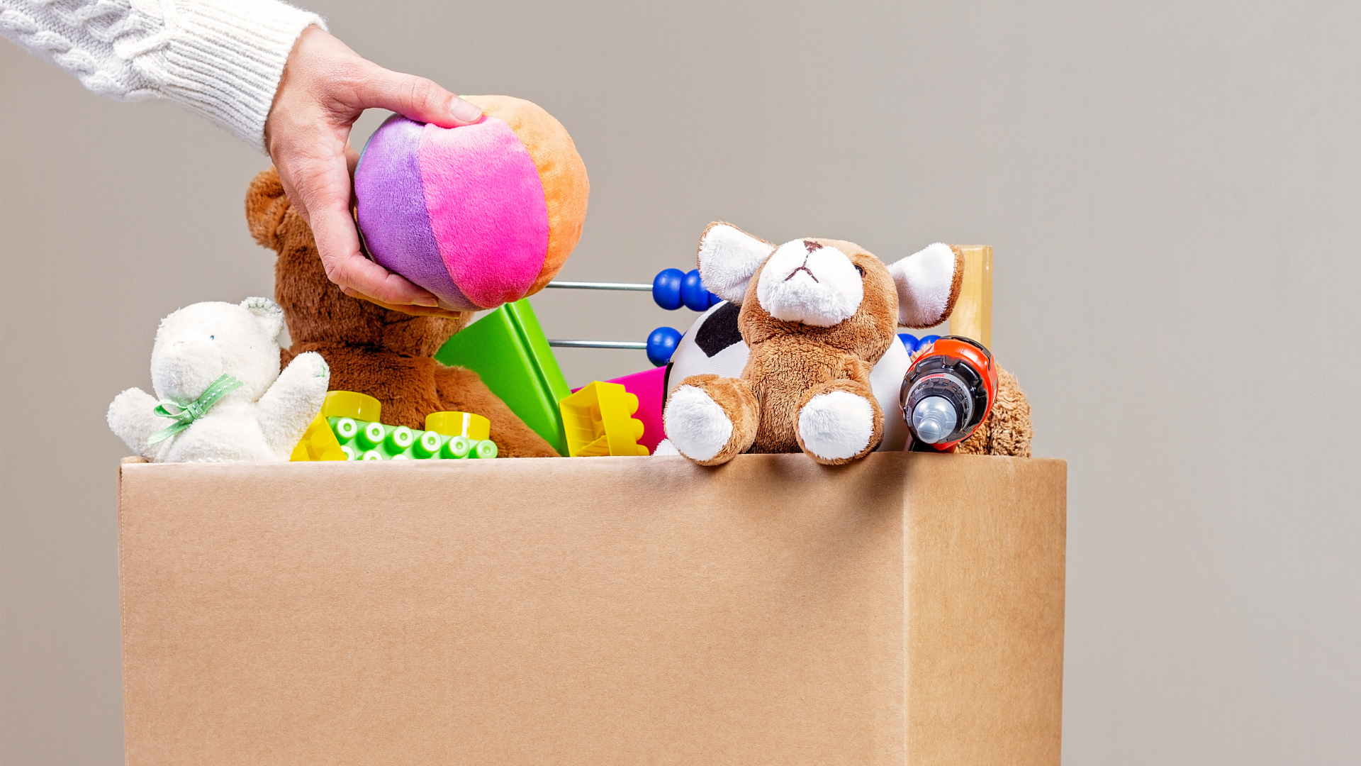 a hand placing a stuffed ball into a cardboard box overflowing with toys