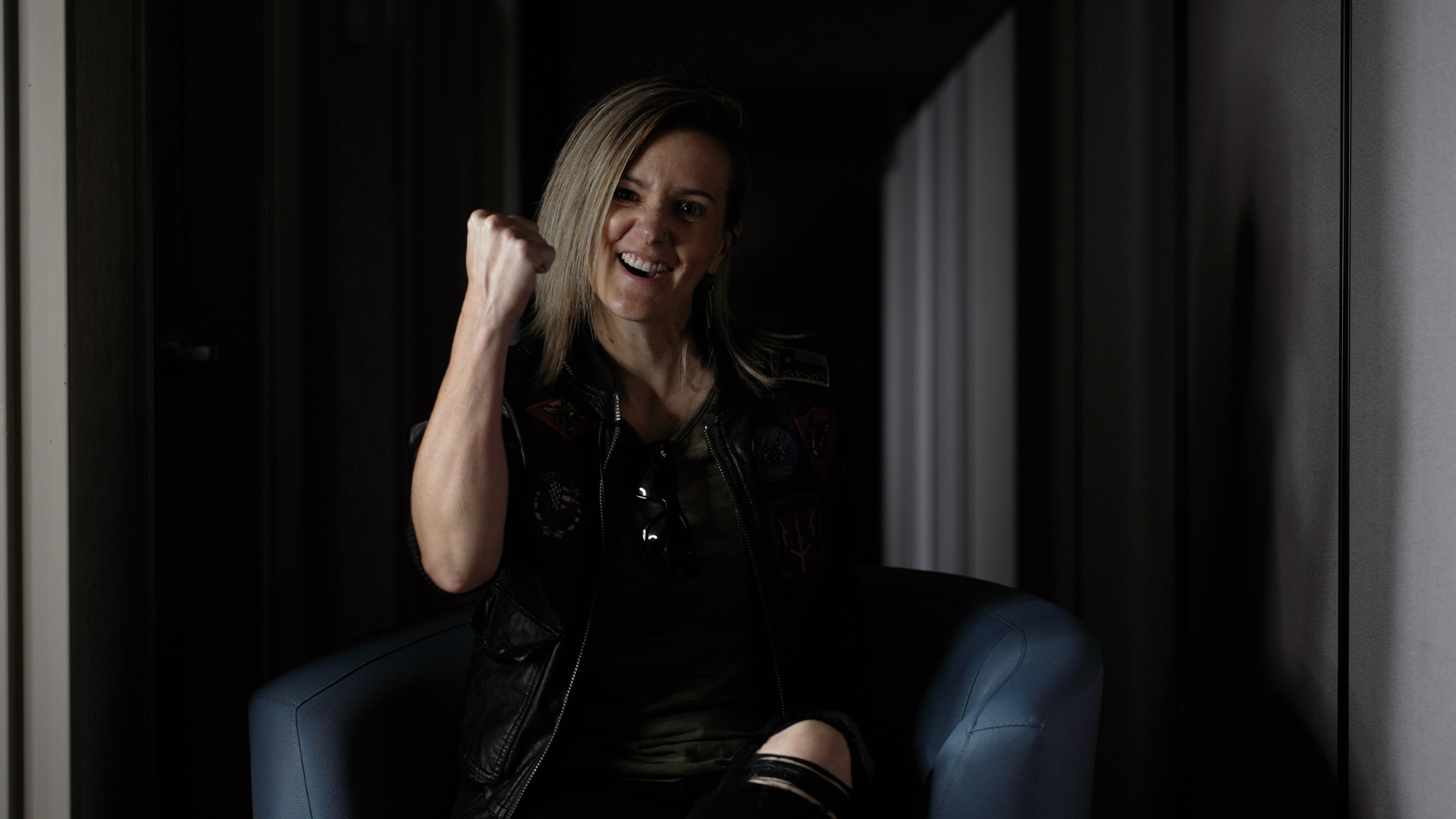 Nicole Hickman sitting in a chair in a dark hallway holding her right arm up and smiling