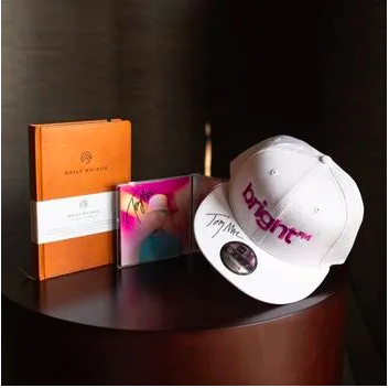 a brown leather Daily Kairos prayer journal, signed TobyMac CD, and signed BRIGHT-FM white snap back hat all on display on a brown table