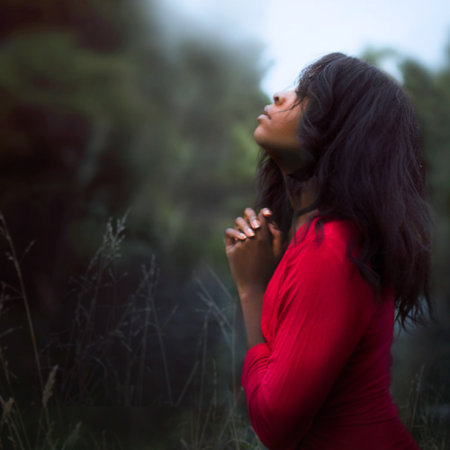 woman wearing a red shirt and kneeling down with her hands folded as she looking toward the sky and prays