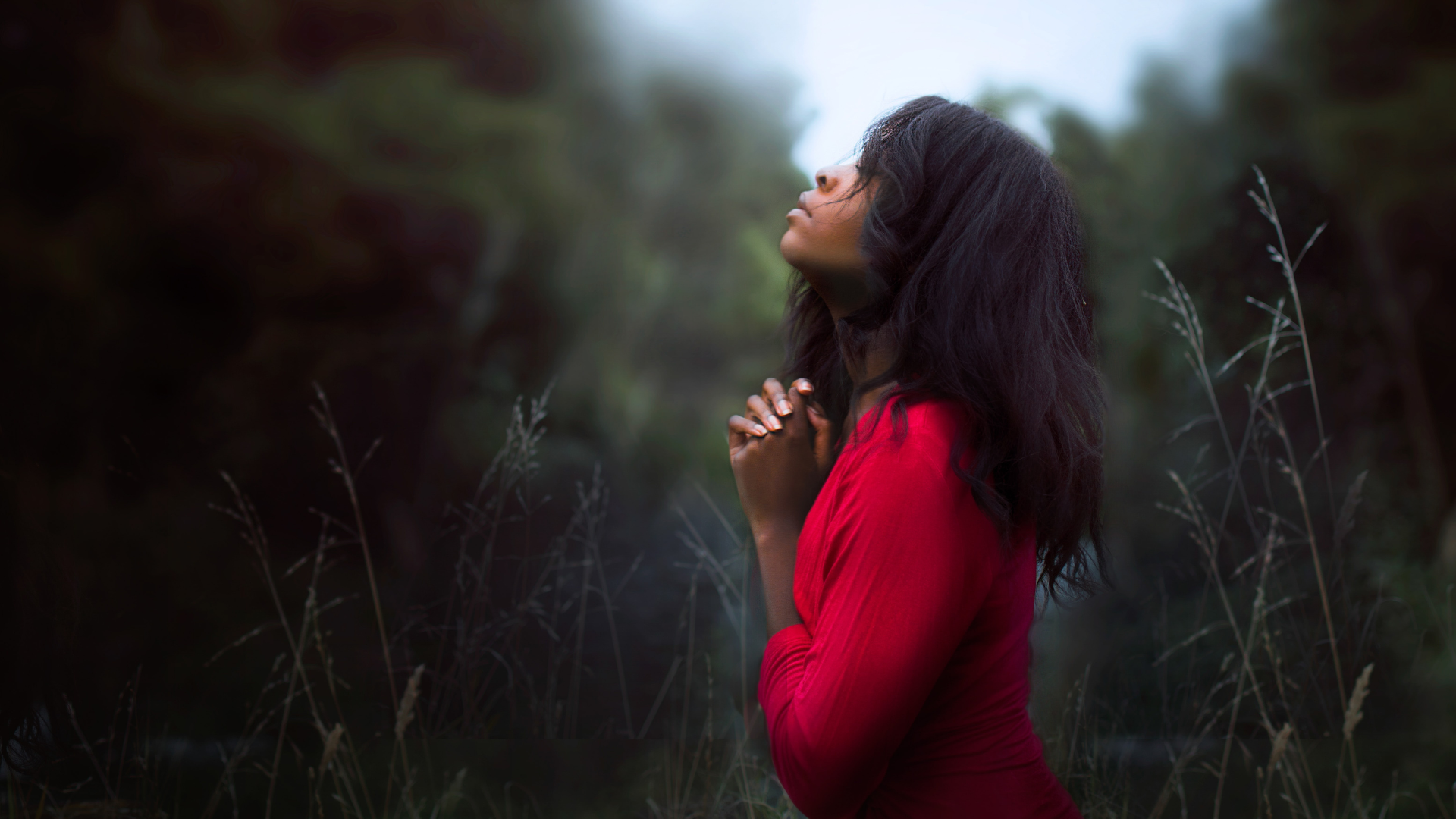 woman wearing a red shirt and kneeling down with her hands folded as she looking toward the sky and prays