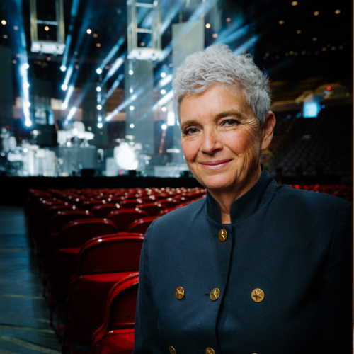 Helen Smallbone sitting in a red seat with rows and rows of red seats behind her in front of a lit up concert stage